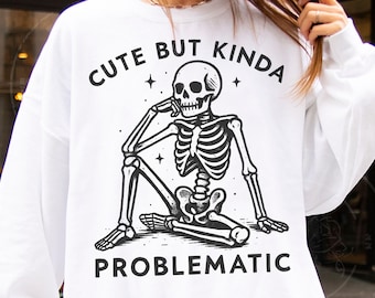 May include: White sweatshirt featuring a black and white illustration of a skeleton in a seated pose. The text "CUTE BUT KINDA PROBLEMATIC" is arched above and below the skeleton. The design includes stars.