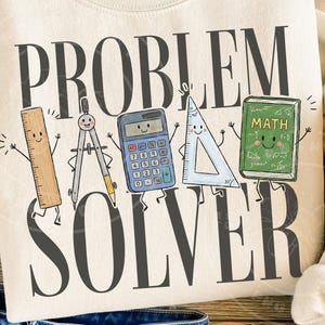Pode incluir: Uma camisola cor de creme com o texto cinzento escuro "PROBLEM SOLVER" e uma ilustração gráfica de cinco artigos escolares sorridentes: uma régua de madeira, um compasso de metal, uma calculadora azul, um esquadro transparente e um livro de matemática verde.