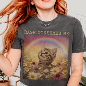 May include: Dark gray t-shirt featuring a graphic of a kitten in a field of flowers under a rainbow. The text "RAGE CONSUMES ME" is printed above the image. The shirt is a relaxed fit.