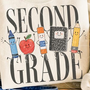 Könnte beinhalten: Ein cremefarbenes Sweatshirt mit dem Text SECOND GRADE in großen dunkelgrauen Buchstaben. Zwischen den Wörtern befinden sich Cartoon-Illustrationen eines blauen Buntstifts, eines roten Apfels, eines orangefarbenen Klebestifts, eines schwarzen Heftes und eines gelben Bleistifts.