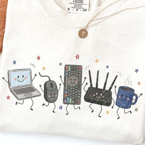 May include: A cream-colored t-shirt featuring a graphic print of five anthropomorphic tech items: a laptop, a computer mouse, a keyboard, a Wi-Fi router, and a blue coffee mug, all with smiling faces and limbs, surrounded by small colorful stars.