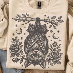 May include: A cream-colored crewneck sweatshirt featuring a black ink-style illustration of a bat hanging upside down from a branch. The bat wears a bow tie and is surrounded by roses, crescent moons, and small stars in a detailed graphic design.
