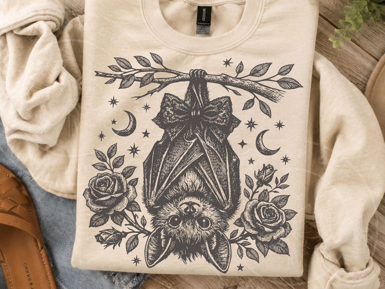 May include: A cream-colored crewneck sweatshirt featuring a black ink-style illustration of a bat hanging upside down from a branch. The bat wears a bow tie and is surrounded by roses, crescent moons, and small stars in a detailed graphic design.