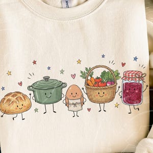 Könnte beinhalten: Ein cremefarbenes Sweatshirt mit Rundhalsausschnitt und einer grafischen Illustration von fünf anthropomorphen Lebensmitteln: ein Brotlaib, ein grüner Topf, ein Ei mit Schürze, ein geflochtener Korb mit Gemüse und ein Glas rote Marmelade mit Sternen und Herzen.