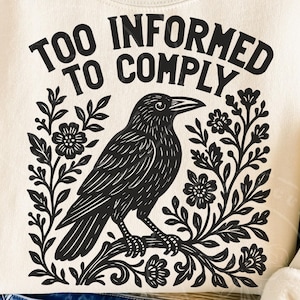Pode incluir: Camisola creme com um desenho gr&aacute;fico preto de um corvo empoleirado num galho, rodeado por elementos florais. O texto "TOO INFORMED TO COMPLY" est&aacute; arqueado acima do corvo. O desenho est&aacute; centrado na frente.