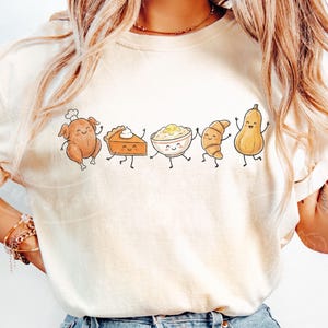 May include: Cream-colored t-shirt featuring a whimsical illustration of Thanksgiving food items. A cartoon turkey, pie, mashed potatoes, croissant, and squash are depicted with smiling faces and dancing poses. The design is centered on the shirt.