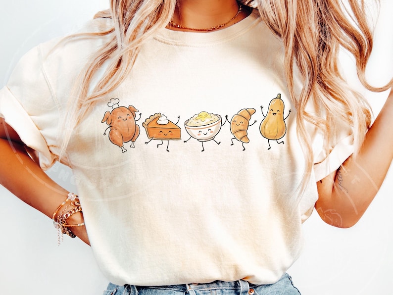 May include: Cream-colored t-shirt featuring a whimsical illustration of Thanksgiving food items. A cartoon turkey, pie, mashed potatoes, croissant, and squash are depicted with smiling faces and dancing poses. The design is centered on the shirt.