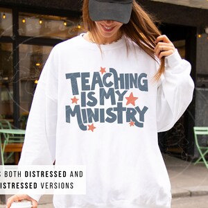 May include: White sweatshirt with the text "TEACHING IS MY MINISTRY" in blue and red. The sweatshirt includes both distressed and non-distressed versions. The image shows a person wearing the sweatshirt.