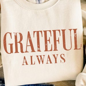 May include: A cream-coloured crewneck sweatshirt featuring the text "GRATEFUL ALWAYS" printed in a distressed, burnt orange font across the chest. The garment has long sleeves and a ribbed neckline, showcasing a simple and clean design.