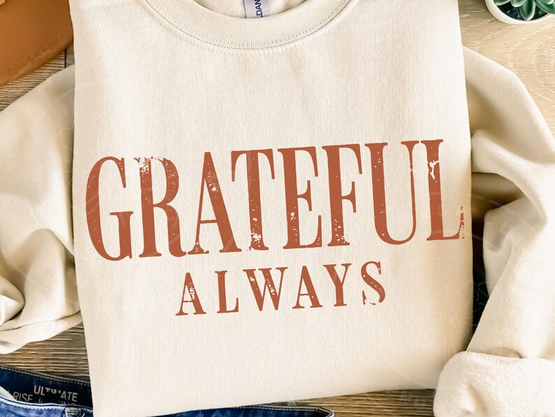 May include: A cream-coloured crewneck sweatshirt featuring the text "GRATEFUL ALWAYS" printed in a distressed, burnt orange font across the chest. The garment has long sleeves and a ribbed neckline, showcasing a simple and clean design.