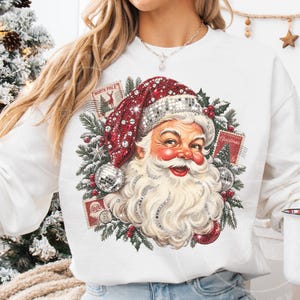 May include: A white long-sleeve sweatshirt featuring a graphic of Santa Claus wearing a red hat with a disco ball texture. The design includes green pine branches, red berries, and vintage-style postage stamps labeled North Pole and Christmas.
