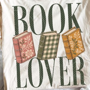 May include: A cream-colored t-shirt featuring the dark green polka-dot text BOOK LOVER. Between the words are three illustrated books: a pink one with floral patterns, a green one with a gingham check, and a yellow one with leaf designs.