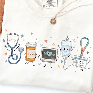 May include: A cream-colored t-shirt featuring a colorful graphic print of five animated medical items: a blue stethoscope, an orange pill bottle, a heart monitor, an IV bag, and an ID badge, all with smiling faces and small colorful stars and hearts.