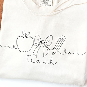 Pode incluir: Camiseta creme com um desenho a traço preto de uma maçã, laço e lápis, com a palavra "Teach" em escrita cursiva. A camiseta tem gola redonda e uma pequena etiqueta na parte superior.