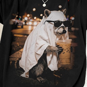 May include: Black t-shirt featuring a raccoon dressed as a ghost, wearing sunglasses, and holding an iced coffee. The raccoon is in front of a blurred city background. The shirt is a dark color.