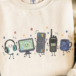 May include: A cream-colored crewneck sweatshirt featuring a graphic print of five anthropomorphic objects: headphones, a computer monitor displaying a map, a keyboard, a two-way radio, and a blue coffee mug, all surrounded by small colorful stars.