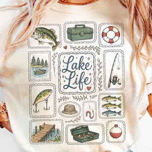 May include: Cream-colored t-shirt featuring a collection of fishing-themed illustrations. The design includes a fish, tackle box, life preserver, fishing rod, and the words "Lake Life". The illustrations are framed in decorative borders.