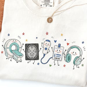 Puede incluir: Una camiseta de color crema con un estampado gráfico de cinco objetos médicos antropomórficos: un escáner CT, una imagen de un cerebro, una sonda de ultrasonido, una tarjeta de identificación y auriculares. Cada objeto tiene una cara sonriente.