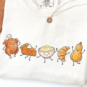 May include: Cream-colored t-shirt featuring a whimsical illustration of Thanksgiving food items. A roasted turkey, pumpkin pie, mashed potatoes, croissant, and butternut squash are depicted with smiling faces and dancing poses. A gold necklace with a pendant is also visible.