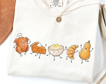 May include: Cream-coloured t-shirt with a playful illustration of Thanksgiving food. A roasted turkey, pumpkin pie, mashed potatoes, croissant, and butternut squash are shown with smiling faces and dancing. A gold necklace with a pendant is also visible.