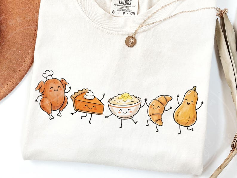 May include: Cream-colored t-shirt featuring a whimsical illustration of Thanksgiving food items. A roasted turkey, pumpkin pie, mashed potatoes, croissant, and butternut squash are depicted with smiling faces and dancing poses. A gold necklace with a pendant is also visible.