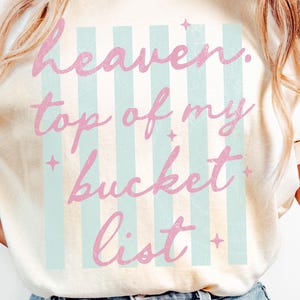 May include: Cream-colored t-shirt with the text "heaven, top of my bucket list" in pink script. The text is set against a backdrop of vertical light blue stripes. Small star graphics are scattered around the text.