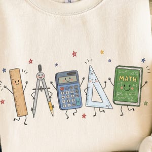 Puede incluir: Una sudadera de color crema con un estampado gr&aacute;fico de cinco &uacute;tiles escolares antropom&oacute;rficos: una regla marr&oacute;n, un comp&aacute;s plateado, una calculadora azul, una escuadra azul claro y un libro verde con la palabra MATH, rodeados de peque&ntilde;as estrellas.
