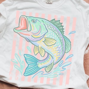 May include: White t-shirt featuring a colorful illustration of a fish jumping out of water. The fish is in shades of blue, green, yellow, and purple, with a pink mouth. Pink and white vertical stripes are behind the fish.