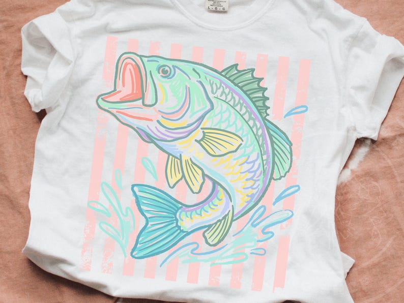 May include: White t-shirt featuring a colorful illustration of a fish jumping out of water. The fish is in shades of blue, green, yellow, and purple, with a pink mouth. Pink and white vertical stripes are behind the fish.
