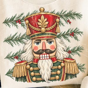 May include: A cream-colored sweatshirt featuring a graphic print of a nutcracker soldier wearing a red and gold uniform with a tall hat. The design includes green pine branches with red berries in the background, framing the central figure.