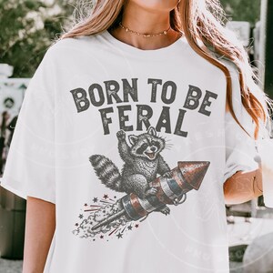 May include: White t-shirt with a graphic of a raccoon riding a rocket, with the text "BORN TO BE FERAL" in gray. The raccoon is in black and white, and the rocket has red, white, and blue stripes.