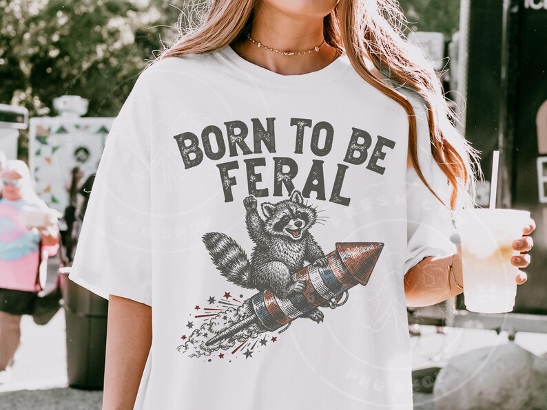 May include: White t-shirt with a graphic of a raccoon riding a rocket, with the text "BORN TO BE FERAL" in gray. The raccoon is in black and white, and the rocket has red, white, and blue stripes.