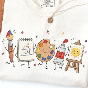 May include: A cream-colored t-shirt featuring a graphic print of five anthropomorphic art supplies including a paintbrush, a sketchbook with a house drawing, a paint palette, a paint tube, and a canvas on an easel, all with smiling faces.