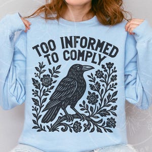 Pode incluir: Camisola azul clara com o texto "TOO INFORMED TO COMPLY" acima de uma ilustra&ccedil;&atilde;o preta de um corvo empoleirado num galho com detalhes florais. O design est&aacute; centrado na frente da camisola.