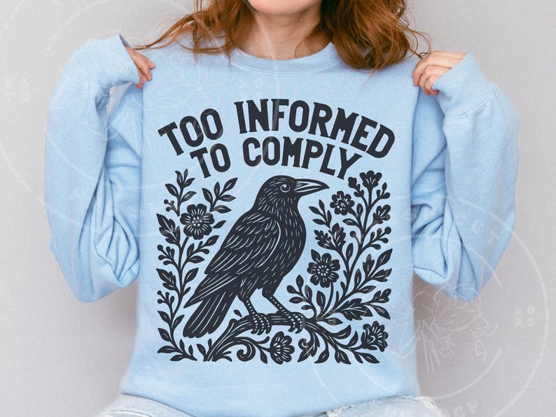 Pode incluir: Camisola azul clara com o texto "TOO INFORMED TO COMPLY" acima de uma ilustra&ccedil;&atilde;o preta de um corvo empoleirado num galho com detalhes florais. O design est&aacute; centrado na frente da camisola.