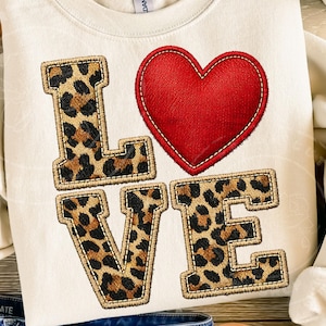 Może przedstawiać: Kremowa bluza z napisem "LOVE". Litery "L", "V" i "E" mają wzór w cętki, a "O" to czerwone serce. Projekt jest wyhaftowany na tkaninie.