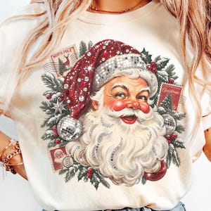 May include: A cream-colored t-shirt featuring a graphic of Santa Claus wearing a red hat with silver disco ball textures. The design includes green pine branches, red berries, and vintage-style postage stamps labeled North Pole and Christmas.