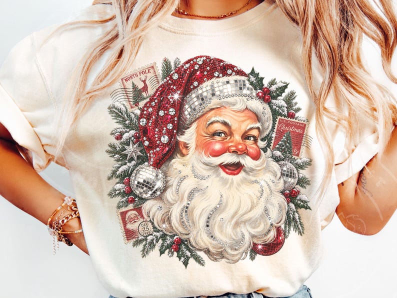 May include: A cream-colored t-shirt featuring a graphic of Santa Claus wearing a red hat with silver disco ball textures. The design includes green pine branches, red berries, and vintage-style postage stamps labeled North Pole and Christmas.
