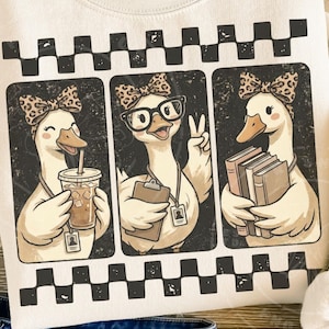 Op de afbeelding: Een crèmekleurig sweatshirt met een grafische print van drie eenden met strikjes met luipaardprint. De eerste eend houdt een ijskoffie vast, de tweede draagt een bril en houdt een klembord vast, en de derde houdt een stapel boeken vast. Een zwart-wit geblokte rand omlijst de boven- en onderkant van het ontwerp.