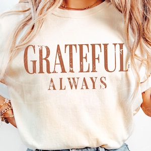 May include: A cream-coloured short-sleeve t-shirt featuring the text "GRATEFUL ALWAYS" printed in a distressed, rust-coloured serif font across the chest. The shirt has a crew neckline and a relaxed fit, showcasing a simple and clean design.