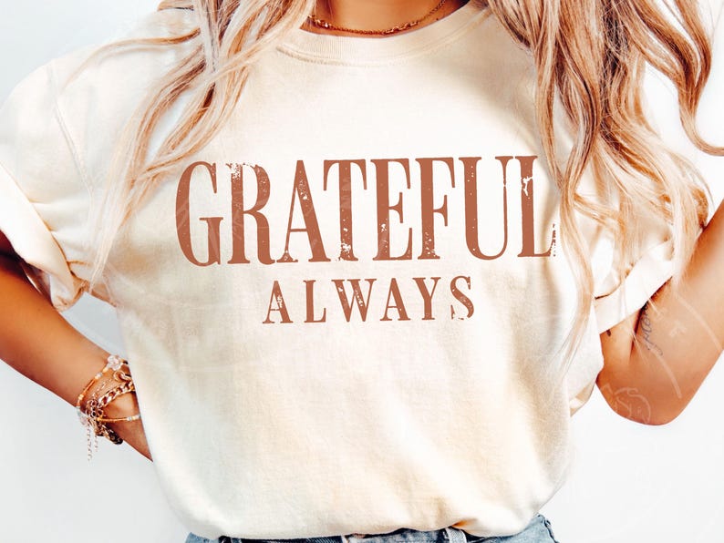 May include: A cream-coloured short-sleeve t-shirt featuring the text "GRATEFUL ALWAYS" printed in a distressed, rust-coloured serif font across the chest. The shirt has a crew neckline and a relaxed fit, showcasing a simple and clean design.