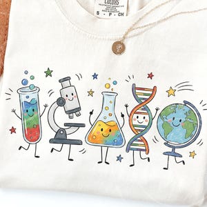 Puede incluir: Una camiseta de color crema con un estampado gráfico colorido de objetos científicos antropomórficos, incluyendo un tubo de ensayo, un microscopio, un matraz Erlenmeyer, una cadena de ADN y un globo terráqueo, todos con caras sonrientes y extremidades negras.