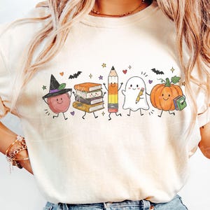 May include: A cream-colored t-shirt featuring a graphic print of a smiling apple wearing a witch hat, a stack of books, a pencil, a ghost holding a pencil, and a pumpkin holding a book, all accented with small stars, hearts, and bats.