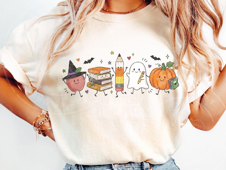 May include: A cream-colored t-shirt featuring a graphic print of a smiling apple wearing a witch hat, a stack of books, a pencil, a ghost holding a pencil, and a pumpkin holding a book, all accented with small stars, hearts, and bats.
