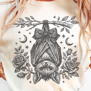 May include: A cream-colored t-shirt featuring a black ink-style graphic of a bat hanging upside down from a branch. The bat wears a bow, surrounded by roses, crescent moons, and small stars. The design is centered on the front of the garment.