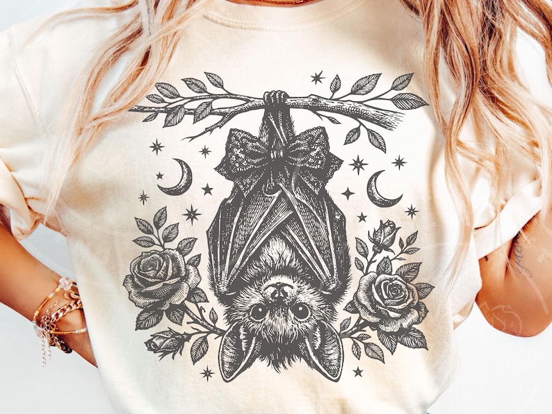 May include: A cream-colored t-shirt featuring a black ink-style graphic of a bat hanging upside down from a branch. The bat wears a bow, surrounded by roses, crescent moons, and small stars. The design is centered on the front of the garment.