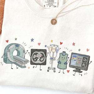 May include: A cream-colored t-shirt featuring a graphic print of cartoon medical imaging equipment, including a CT scanner, a cross-section scan, an IV pole, a lead apron, and a computer monitor, all with smiling faces, surrounded by small stars and hearts.