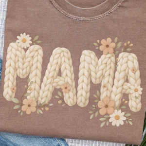 May include: Brown t-shirt with the word "MAMA" in a cream-colored, knitted font, adorned with floral accents. The flowers are in shades of cream and peach, with green leaves. The shirt is folded on a white surface with blue jeans.