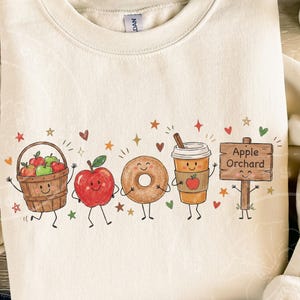 May include: A cream-colored crewneck sweatshirt featuring a graphic print of a smiling apple basket, a red apple, a donut, a coffee cup, and a wooden sign that reads Apple Orchard, all surrounded by small colorful stars and hearts.