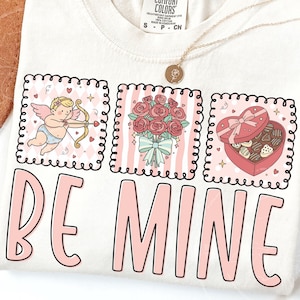 Könnte beinhalten: Cremefarbenes T-Shirt mit Valentinsthema. Das Shirt zeigt drei illustrierte Quadrate mit einem Amor, einem Rosenstrauß und einer herzförmigen Pralinenschachtel. Die Worte "BE MINE" sind in Rosa gedruckt.
